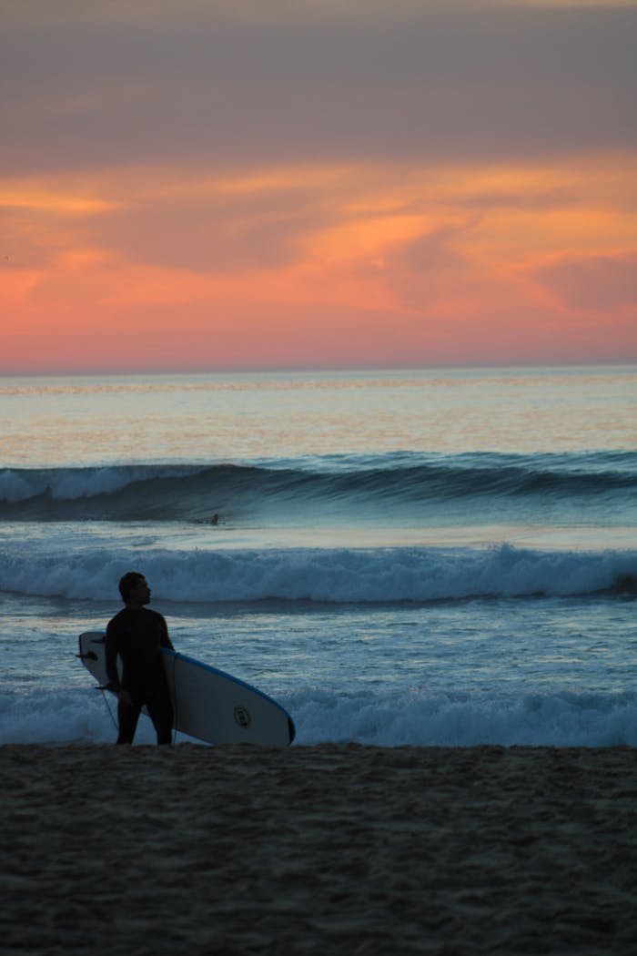 A surfer standing at the shoreline, watching the sunset over the waves in Torres Vedras, Portugal.
